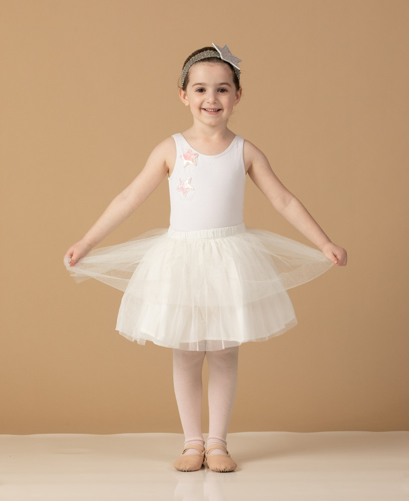 Young dancer aged 3 to 5 wearing standing upright and smiling in a cute white dress. Holding out the skirt to the sides. Metrowest Dance Academy professional photo shoot.