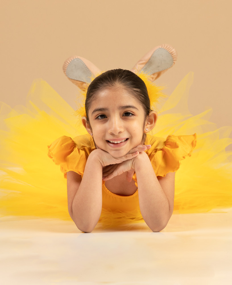 Young dancer aged 5 to 8 wearing a bright yellow dress, lying down with head resting on hands and smiling at the camera. Metrowest Dance Academy professional photo shoot.